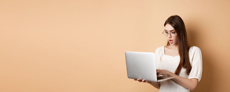 Worried girl look at laptop screen, typing on computer with focused face. Woman freelancer working in internet, standing on beige background.