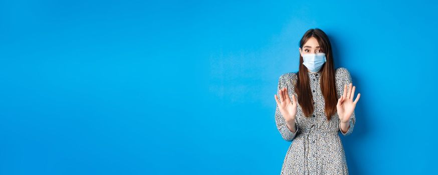 Covid-19, pandemic lifestyle concept. No thank you. Reluctant and worried girl in medical mask asking non-masker to stay away, showing block gesture, tell stop, blue background.