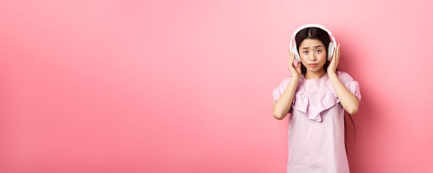 Skeptical girl unamused with song, listening music in headphones and frowning displeased, standing against pink background.