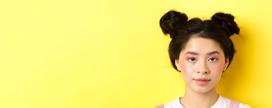 Head portrait of stylish asian girl with bright makeup and hairbuns, looking at camera, standing on yellow background.