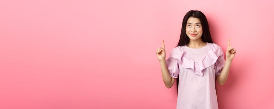 Beautiful smiling asian teen girl pointing fingers up, looking at advertisement with happy face, standing on pink background.