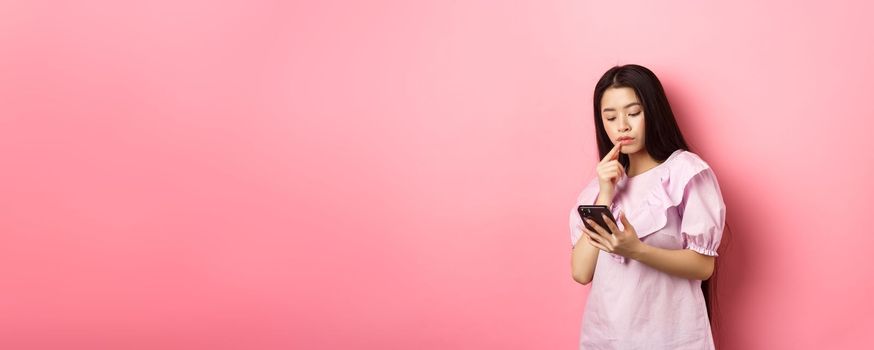 Online shopping. Pensive asian girl looking at smartphone screen, making choice, standing in dress against pink background.