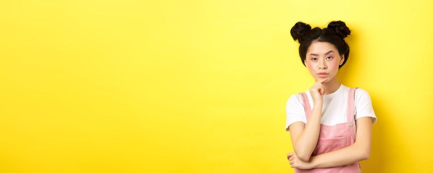 Suspicious asian girl looking at camera and thinking, stare with disbelief, standing with makeup on yellow background.