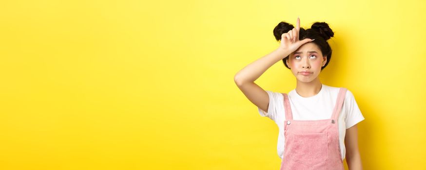 Sad girl showing loser sign on forehead and sulking upset, feeling disappointed in herself, standing on yellow background.