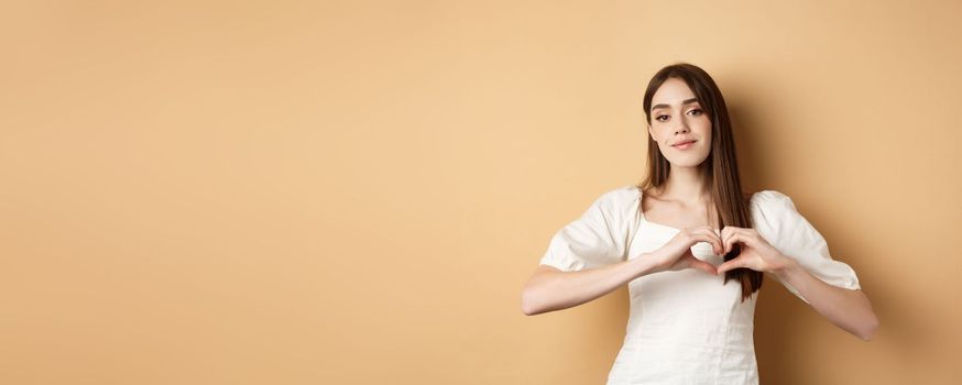 Valentines day and romance concept. Romantic caucasian girl in white dress showing heart gesture and smiling, say I love you, standing over beige background.