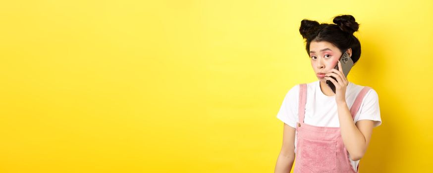 Cute teen girl talking on smartphone, making silly pouting face and look timid at camera, standing with glamour makeup on yellow background.