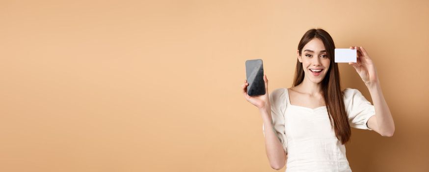 Cheerful young woman showing plastic credit card and empty mobile phone screen, smiling pleased at camera, standing on beige background.