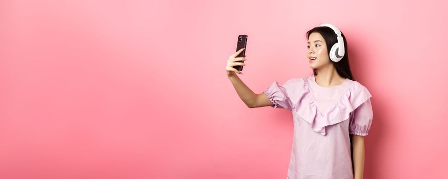 Beautiful asian girl blogger taking selfie in wireless headphones, make photo for social media on smartphone, standing in dress against pink background.