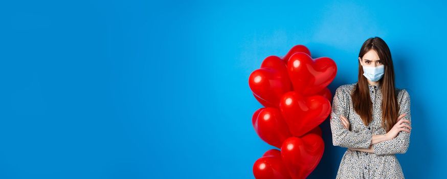 Covid-19, quarantine and health conept. Angry girlfriend in medical mask, standing near Valentines day red heart balloons, cross arms on chest and frowning offended, blue background.