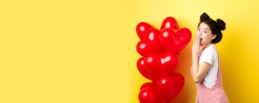 Valentines day and relationship concept. Coquettish and romantic girl laughing, covering mouth with hand, look silly at camera, standing near red heart balloons, yellow background.