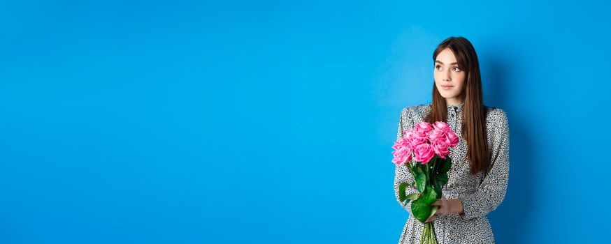 Valentines day concept. Pretty romantic girl looking dreamy at empty space, holding bouquet of pink roses on date, standing alone on blue background.
