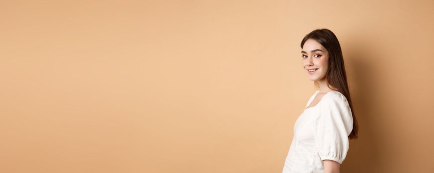 Profile of pretty european girl turn head at camera and smiling coquettish, standing in white blouse against beige background.