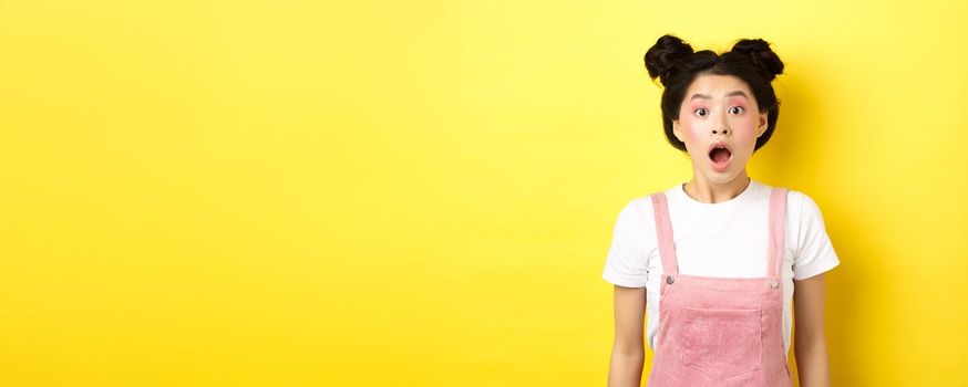 Excited asian teen girl drop jaw, gasping amazed with opened mouth, looking at camera impressed, checking out summer promo offer, standing on yellow background.