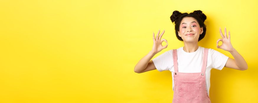 Happy teen koren girl with beauty makeup, showing okay signs and smiling satisfied, recommending product, praise good choice, standing on yellow background.