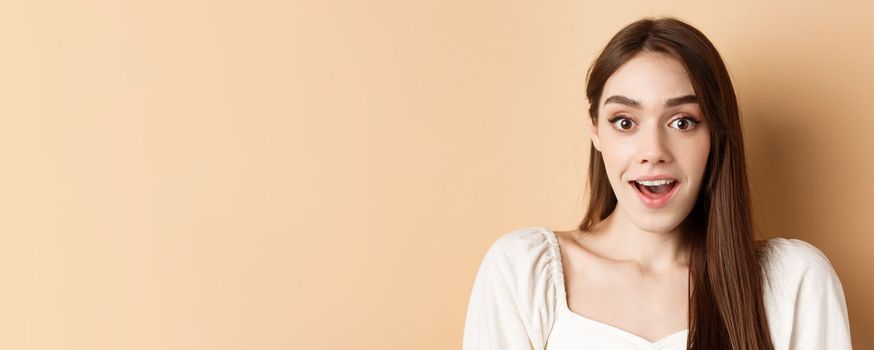 Excited young woman gasping amazed, saying wow and looking at camera, checking out promo offer, standing on beige background.