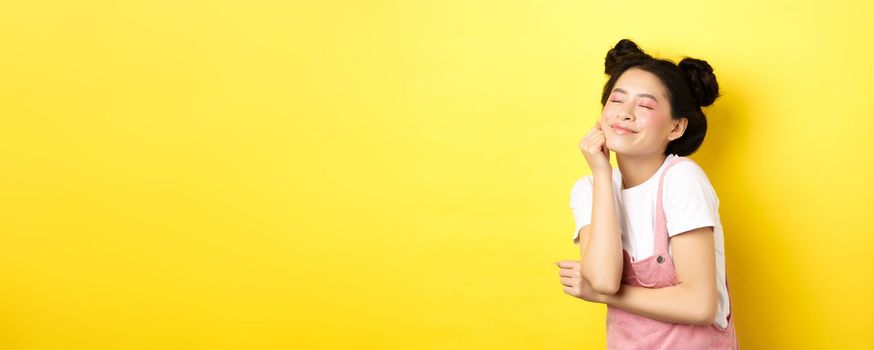 Beautiful happy woman with bright pink makeup, close eyes and touch soft skin, smiling excited, standing in summer clothes on yellow background.