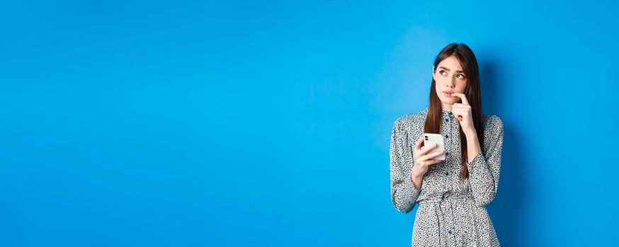 Pensive cute girl thinking how to answer on message, looking aside thoughtful and holding smartphone, standing in dress on blue background.