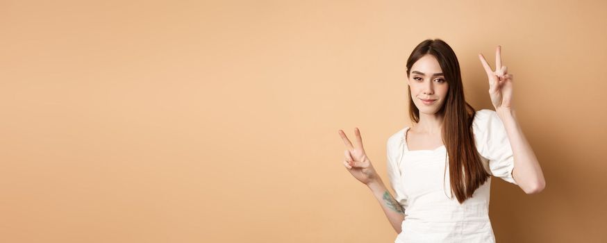 Cheerful attractive girl dancing and having fun, showing v-signs and looking positive at camera, standing on beige background.