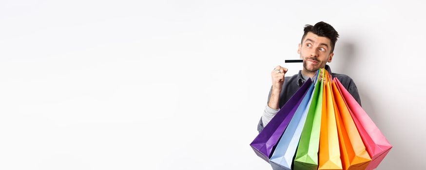 Dreamy guy thinking of shopping, holding colorful bags with purchased items and standing thoughtful with credit card, white background.