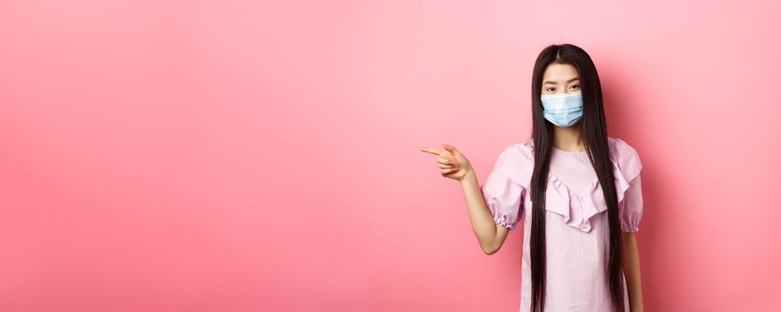 Covid-19, pandemic lifestyle concept. Cute asian teen girl in medical mask pointing finger left at logo, showing advertisement, standing in dress on pink background.