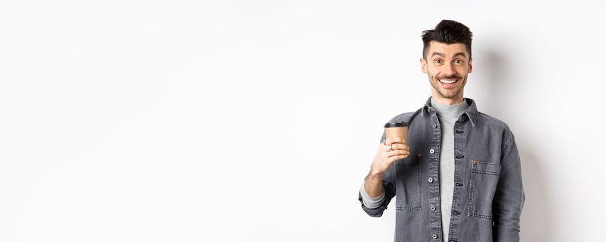 Enthusiastic handsome man holding coffee cup and smiling, drinking good drink from cafe takeout, standing against white background.