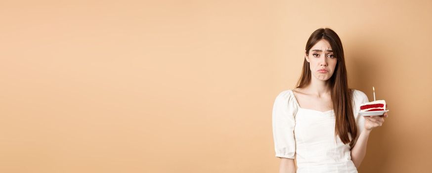 Sad birthday girl holding cake with candle and look upset, feeling lonely on her bday, standing alone on beige background.