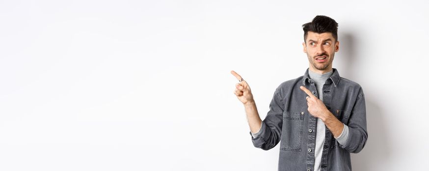 Skeptical and doubtful young man pointing fingers left at logo, look with dislike and confusion, standing on white background.
