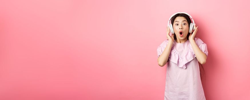 Excited asian girl hear awesome song, listen music in headphones and look amazed, standing on pink background.