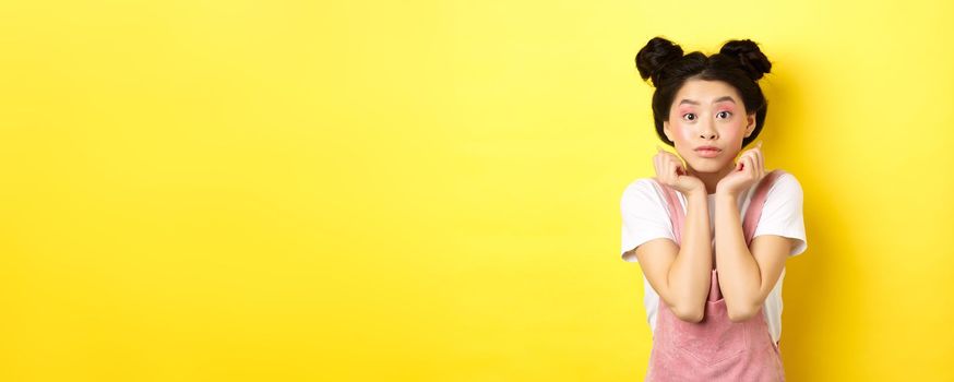Cute teenage asian girl with beauty makeup, listen with interest, lean on hands and looking excited at camera, standing on yellow background.