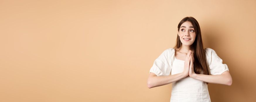 Hopeful young girl begging god and looking dreamy at upper left corner, praying or making wish, standing on beige background.