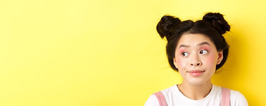 Close-up portrait of cute glamour asian girl, bright makeup and hairstyle, looking aside at logo with silly face, yellow background.
