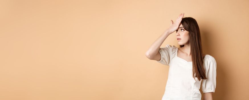 Girl rolling her eyes and slap forehead annoyed, bothered by something stupid or lame, making face palm on beige background.