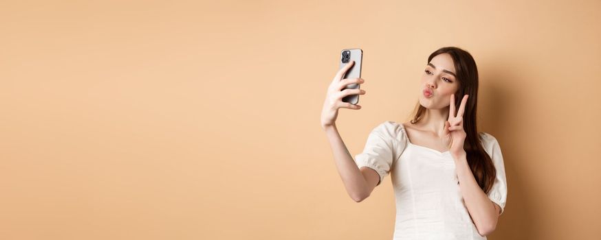 Fashion female blogger pucker lips and show v-sign at smartphone camera, taking selfie for social media, standing on beige background.