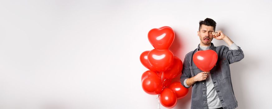 Valentines day and love concept. Sad crying man holding red heart balloon and whiping tears, standing single and miserable, being heartbroken, white background.