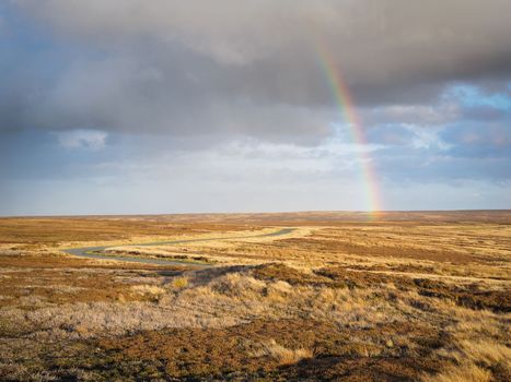 Rainbow arcs over the winding road and moorland under moody sky and dark clouds at the head of the Rosedale valley, North York Moors National Park, UK