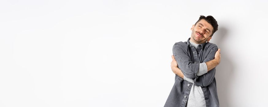 Happy romantic guy hugging himself with closed eyes and smile, loving own body, daydreaming about lover, standing on white background.