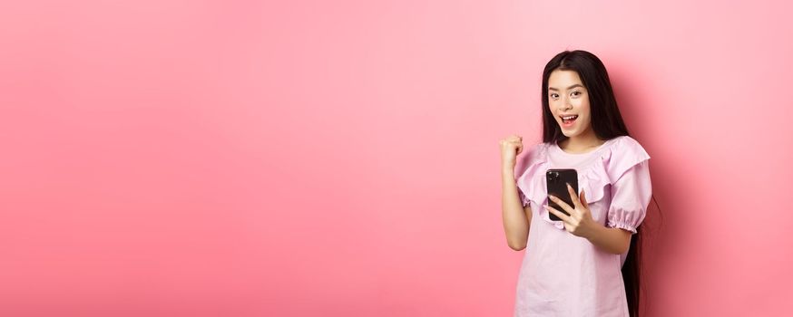 Online shopping. Satisfied asian girl winning on mobile phone, say yes and make fist pump, holding smartphone, standing against pink background.