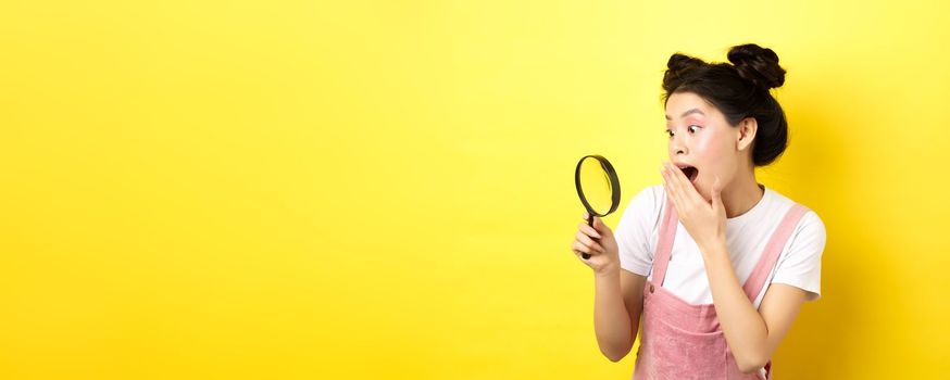 Excited silly asian girl gasping wondered, found something interesting, looking through magnifying glass, standing on yellow background.