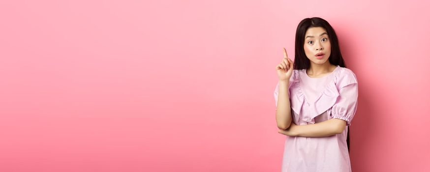 Excited asian woman pitching an idea, raising finger in eureka sign, have plan or solution, standing on pink background.