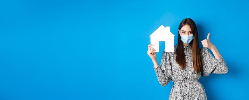 Real estate, covid-19 and pandemic concept. Candid woman in medical mask showing thumb up and paper house cutout, reanting apartment, standing on blue background.
