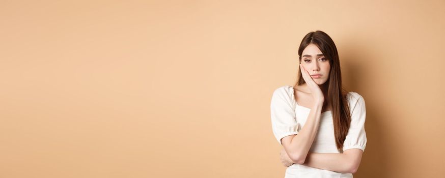 Bored and tired young woman looking indifferent at camera, lean face on palm and stare at camera unamused, standing sleepy on beige background.