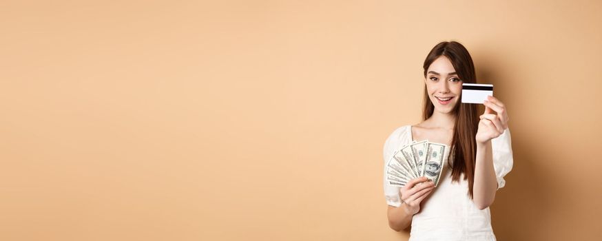 Smiling girl showing platic credit card to camera, holding dollar bills close to chest, standing on beige background.