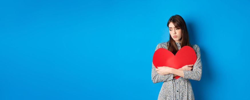 Valentines day. Lonely single girl hugging big red heart cutout and looking down upset, standing gloomy on blue background.