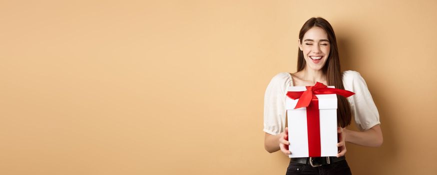 Happy smiling woman laughing, holding gift box from lover, enjoying romantic date on Valentines day, standing on beige background.