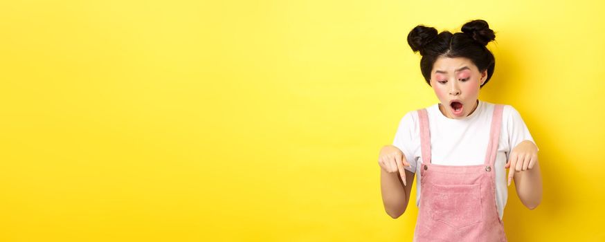 Excited teenage asian girl checking out advertisement, pointing fingers down and drop jaw amazed, showing promo offer, yellow background.