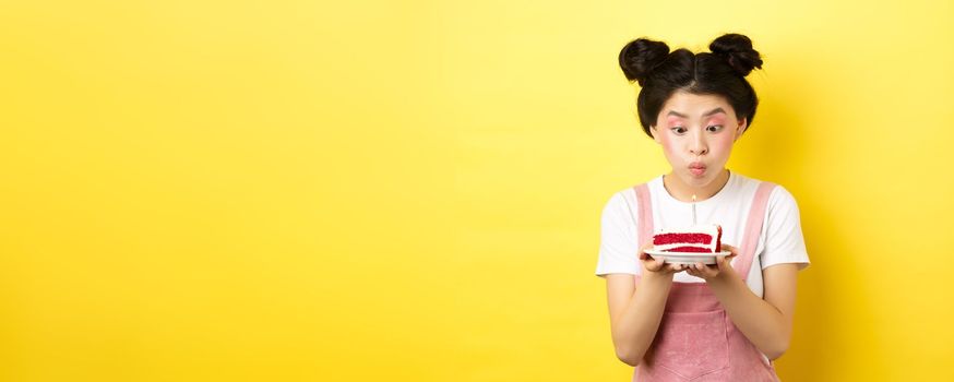 Holidays and celebration. Silly asian girl with glamour makeup, making wish and blowing candle on birthday cake, standing on yellow background.