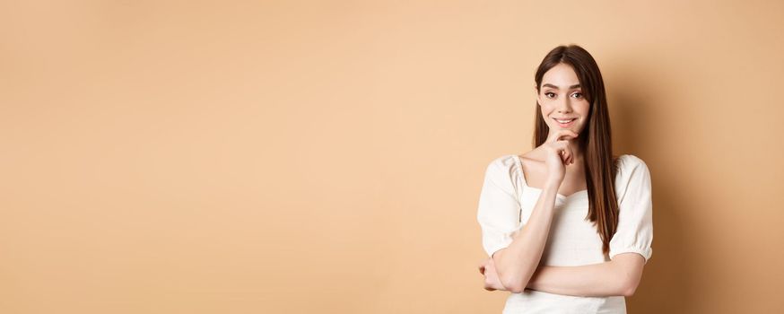 Smiling young european woman in dress, having an idea, listening with interest, touching chin and looking at camera intrigued, standing against beige background.