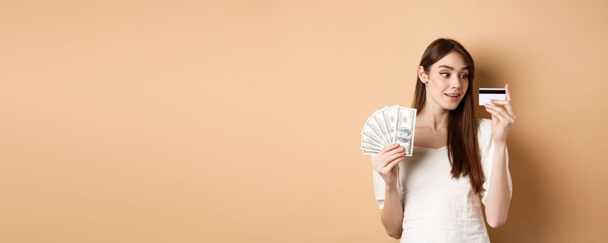 Dreamy girl looking at plastic credit card and thinking of shopping, holding dollar bills, standing on beige background.