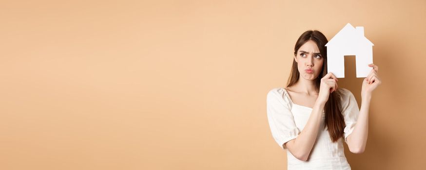 Real estate. Upset young woman showing paper house cutout and frowning sad, looking aside thoughtful, thinking of buying apartment, standing on beige background.