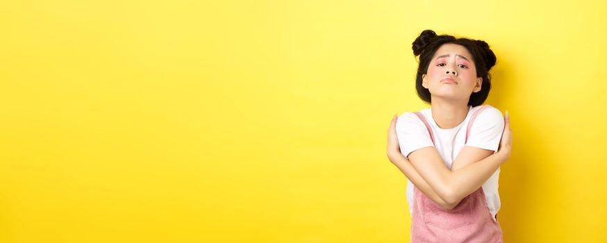 Lonely sad teen girl hugging herself, wanting to relationship and cuddles, standing alone on yellow background.